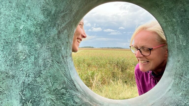 Two people smiling through a circular opening in a sculpture, with a field and cloudy sky in the background